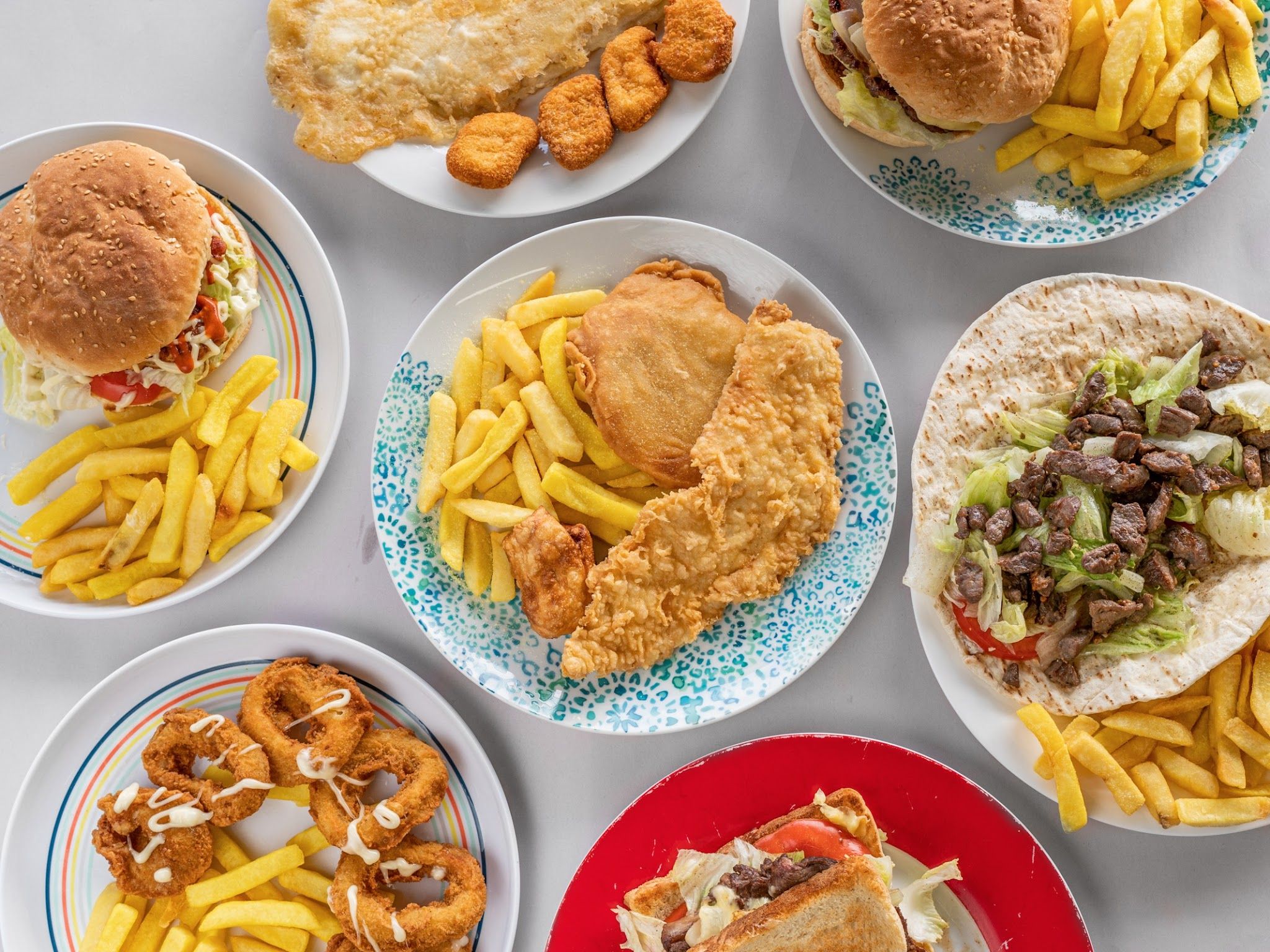 Wide overhead flat-lay of seven plates including battered fish, chips, burgers, calamari and wraps